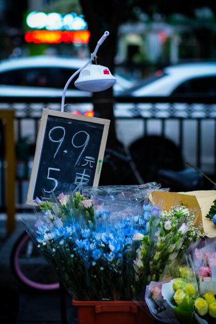 A display of fresh floral bouquets situated outdoors, featuring arrangements of pink, white, and yellow flowers wrapped in clear plastic. The bouquets are arranged in baskets and cardboard boxes on a 