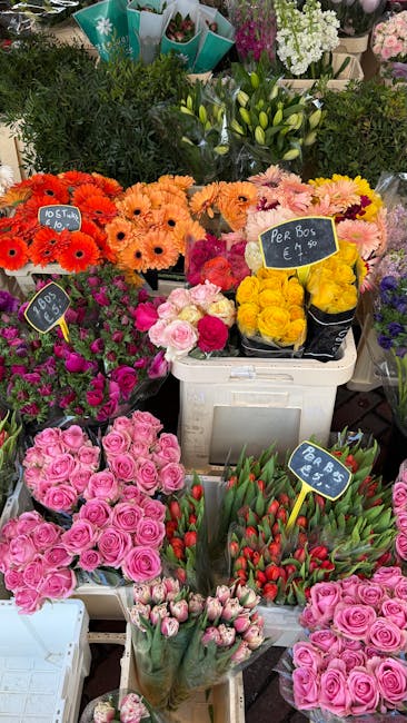 A vibrant display of fresh flowers arranged in various containers at a flower shop, including pink roses, tulips, yellow roses, orange gerbera daisies, and pink carnations. The flowers are organized i