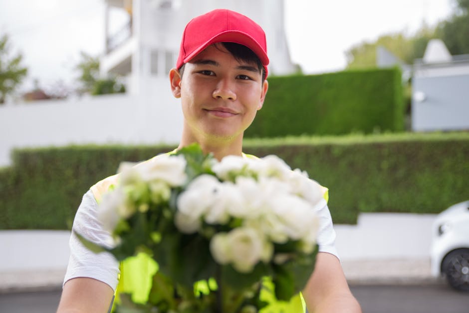 A delivery person wearing a red cap and high-visibility vest holding a bouquet of fresh white roses and green foliage, extending it towards the camera. The bouquet is arranged with lush, vibrant petal