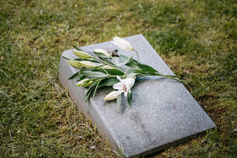 A bouquet of fresh white lilies with large, open petals and long green leaves rests atop a gray granite memorial stone on a grassy area. The lilies are arranged in a loose, natural style, with some bu