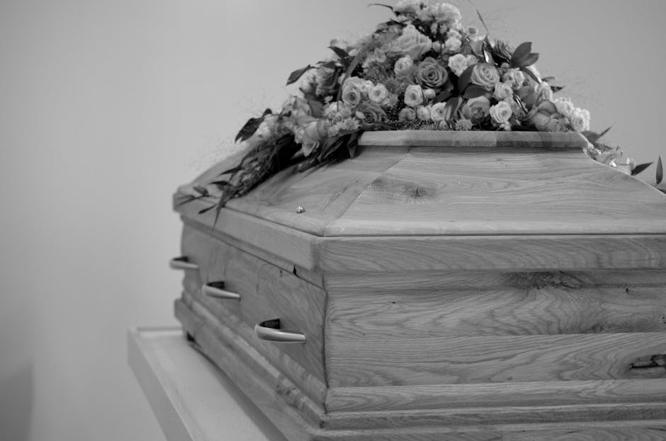 A wooden coffin with smooth, polished surface and visible natural grain, resting on a stand against a plain background. The coffin is adorned with a large, colorful funeral flower arrangement on top, 