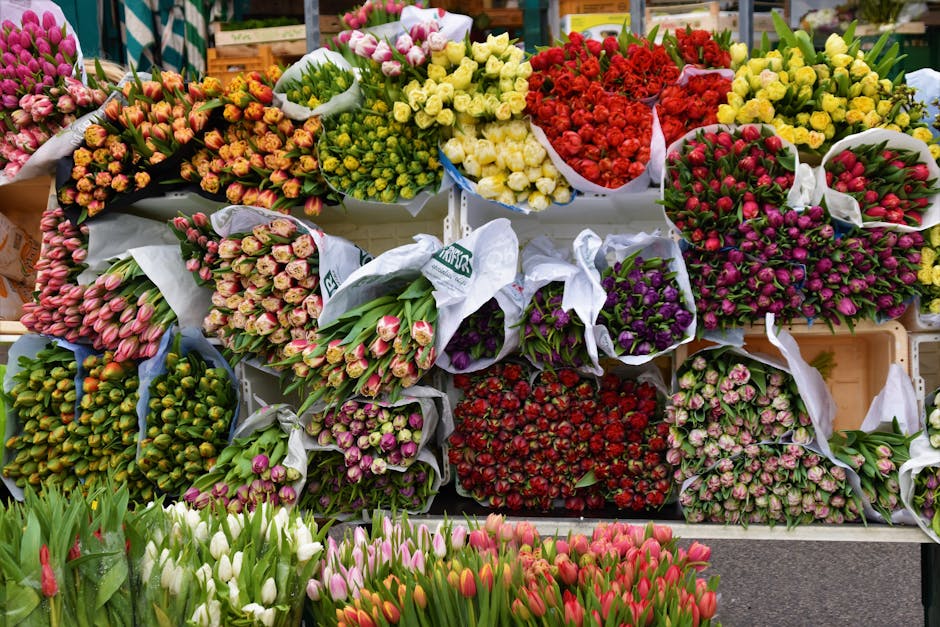 An array of fresh flower bouquets and bunches displayed on a market stand, including pink, yellow, red, purple, and white blooms such as tulips, roses, and ranunculus, wrapped in white and patterned p