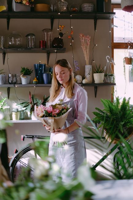 A young woman with long, light brown hair, dressed in a white shirt and apron, holds a bouquet of fresh pink and cream flowers wrapped in brown paper. She stands in a well-lit indoor space with natura