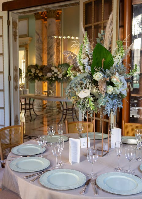 A table setting at an elegant event featuring a tall, lush floral arrangement with white roses, large green leaves, wispy pampas grass, and silver-toned foliage, placed in a copper-colored geometric s