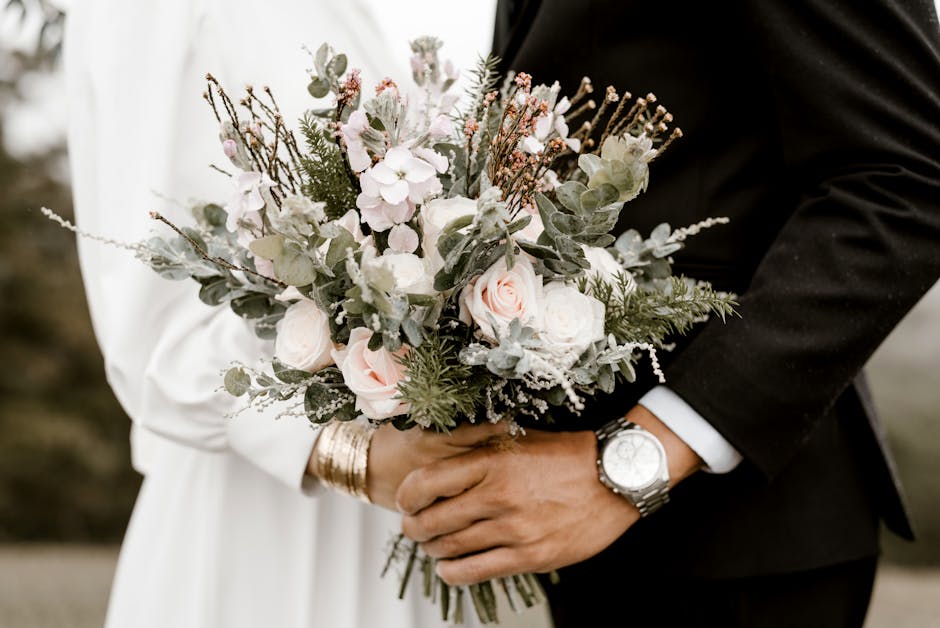 A close-up of a bridal bouquet held by a bride and groom during a wedding ceremony. The bouquet features soft pink roses, white peonies, and delicate sprigs of lavender, along with green eucalyptus le